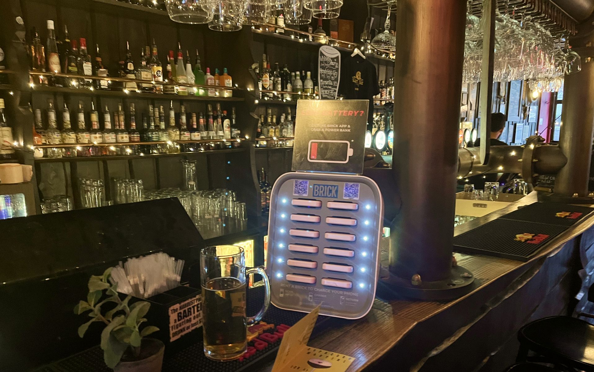 The grey 12 slot Brick powerbank sharing station on the bar at Crazy Horse with a shelf of bottles in the background